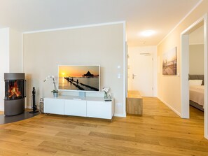 Un salon moderne avec une cheminée, un téléviseur à écran plat fixé au mur, un plancher en bois et un couloir menant à une chambre à coucher.