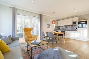 Un salon moderne comprenant un canapé, un fauteuil, une table à manger et un coin cuisine avec des placards et des appareils électroménagers.