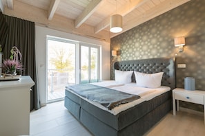 A bedroom with a large bed, a nightstand, a lamp, and a view through a sliding glass door. - Domicile Düne 8, App. 7 (Heringsdorf)