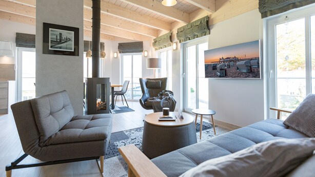 A modern living room with a sofa, armchair, and round coffee table. A fireplace is visible in the background, and there are large windows with a view of the outside. - Domicile Düne 8, App. 7 (Heringsdorf)