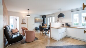 Eine moderne Küche mit weißen Schränken, ein Essbereich mit einem Holztisch und -stühlen sowie ein Wohnbereich mit einem schwarzen Ledersofa und einem rosa Hocker.