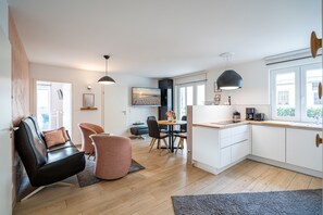 Une cuisine moderne avec des meubles blancs, un coin repas avec une table et des chaises en bois, et un salon avec un canapé en cuir noir et un pouf rose.