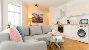 A modern living room with a sofa, a glass coffee table, and a kitchen area with white cabinets and appliances. - Quartier am Kurpark, app. 08 (Heringsdorf)