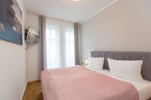 Une chambre à coucher avec un lit, une fenêtre avec des rideaux, un téléviseur fixé au mur et un tableau.