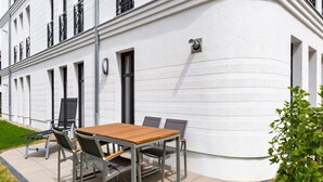 Eine Terrasse im Freien mit einem Holztisch und Metallstühlen, ein weißes Gebäude mit schwarzen Fensterrahmen und eine kleine, grüne Pflanze.