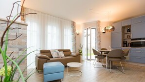 A modern living room with a sofa, ottoman, and coffee table, a dining area with chairs, and a kitchen with cabinets and appliances. - Quartier am Kurpark, app. 02 (Heringsdorf)