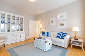 A living room with a white sofa, blue and white cushions, a white cabinet with glass doors, and a wicker coffee table. - House with a view of the sea, Apt. 8 (Heringsdorf)