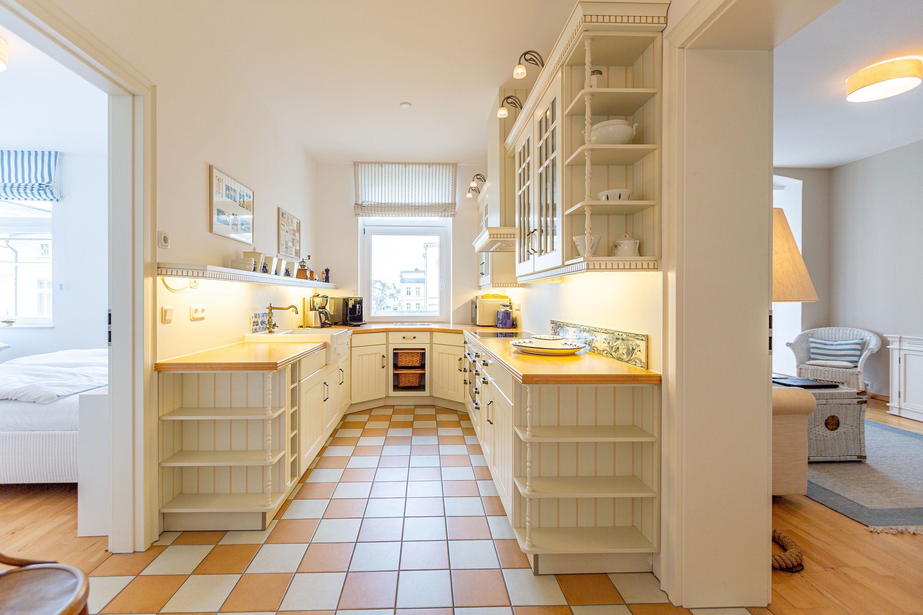 Une cuisine avec des meubles blancs, un carrelage à motifs et une fenêtre avec des stores.