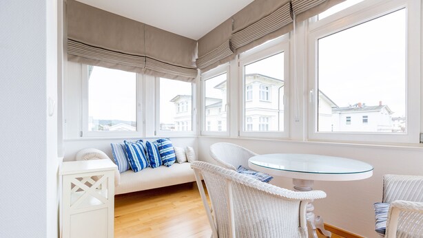 Una habitación luminosa con un sofá blanco, almohadones a rayas, sillas de mimbre, una mesa de vidrio y grandes ventanales con vistas a edificios.