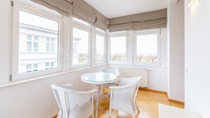 Una habitación luminosa con una mesa redonda de cristal y sillas de mimbre blanco, ventanas grandes y un radiador blanco.