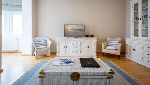 Una sala de estar con un televisor sobre un mueble blanco, dos sillones de mimbre con cojines a rayas, un tapete azul y blanco a rayas, y una estantería con cajas de diseño azul y blanco.