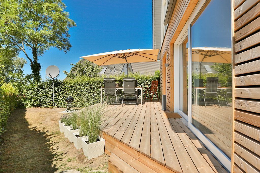 A wooden deck with outdoor furniture and a large umbrella, surrounded by a garden and a house with a glass door.