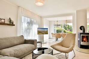 A modern living room with a sofa, a chair, a TV, a fireplace, and a dining area with a view of greenery. - Vacation cottage Hygge Hus (Sellin/Usedom)