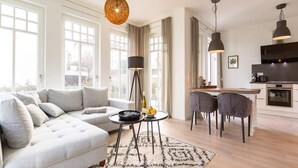 Un salon moderne avec un canapé, une table basse et un coin repas avec des chaises. Il y a de grandes fenêtres, une cuisine avec une cuisinière et des luminaires suspendus.