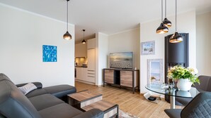 En moderne stue med sofa, sofabord og spiseområde med stole. Der er et køkken i baggrunden med skabe og et fjernsyn.