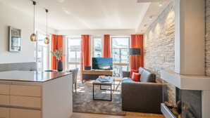 A modern living room with a sofa, coffee table, and TV. There is a kitchen area with a fridge and a dining table. The room has large windows with curtains and a stone wall. - Beachhouse Bansin, app. 22 (Heringsdorf)