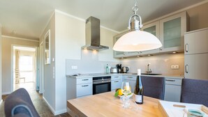 A modern kitchen with a wooden dining table, a hanging light fixture, and a view of the adjacent living area. - Quartier am Fischerstrand, App. 3 (Heringsdorf)