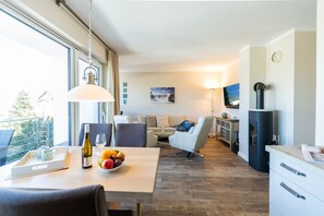 A modern living room with a dining area, a sofa set, a TV, a kitchen area, and a large window.