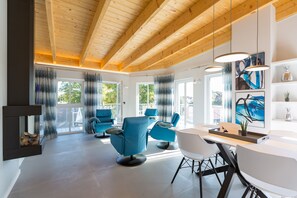 Un interior moderno con vigas de madera, ventanas amplias, sillas azules, una mesa blanca y arte abstracto.
