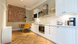 A modern kitchen with white cabinets, a herringbone wood floor, a brick wall, and a dining area with a yellow chair. - Villa Bellevue, App. 03 (Heringsdorf)