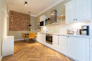 A modern kitchen with white cabinets, a herringbone wood floor, a brick wall, and a dining area with a yellow chair.