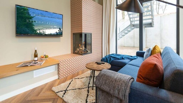 A modern living room with a fireplace, a large TV, a sofa, and a window with a view of a staircase. - Villa Bellevue, App. 03 (Heringsdorf)