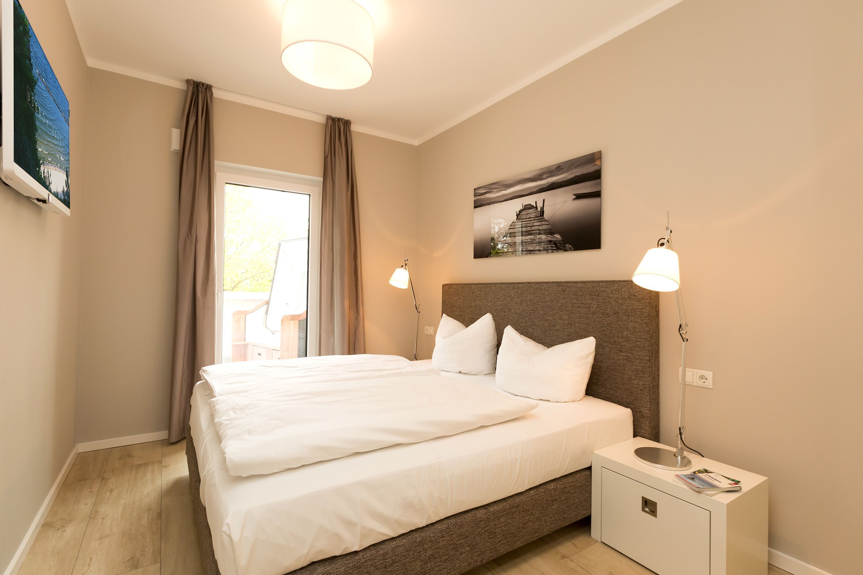 Een slaapkamer met een bed, nachtlampen, een nachtkastje, een raam met gordijnen en een aan de muur hangend schilderij.