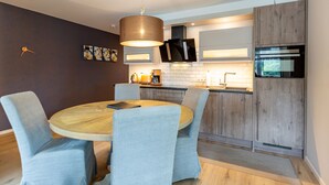 A modern kitchen with a round wooden table and grey upholstered chairs, a built-in oven, and a hanging light fixture. - Dune Residence Heringsdorf, App. 17 (Heringsdorf)