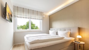 A bedroom with a bed, a bedside table with a lamp, a window with curtains, and a wall-mounted TV. - Dune Residence Heringsdorf, App. 17 (Heringsdorf)