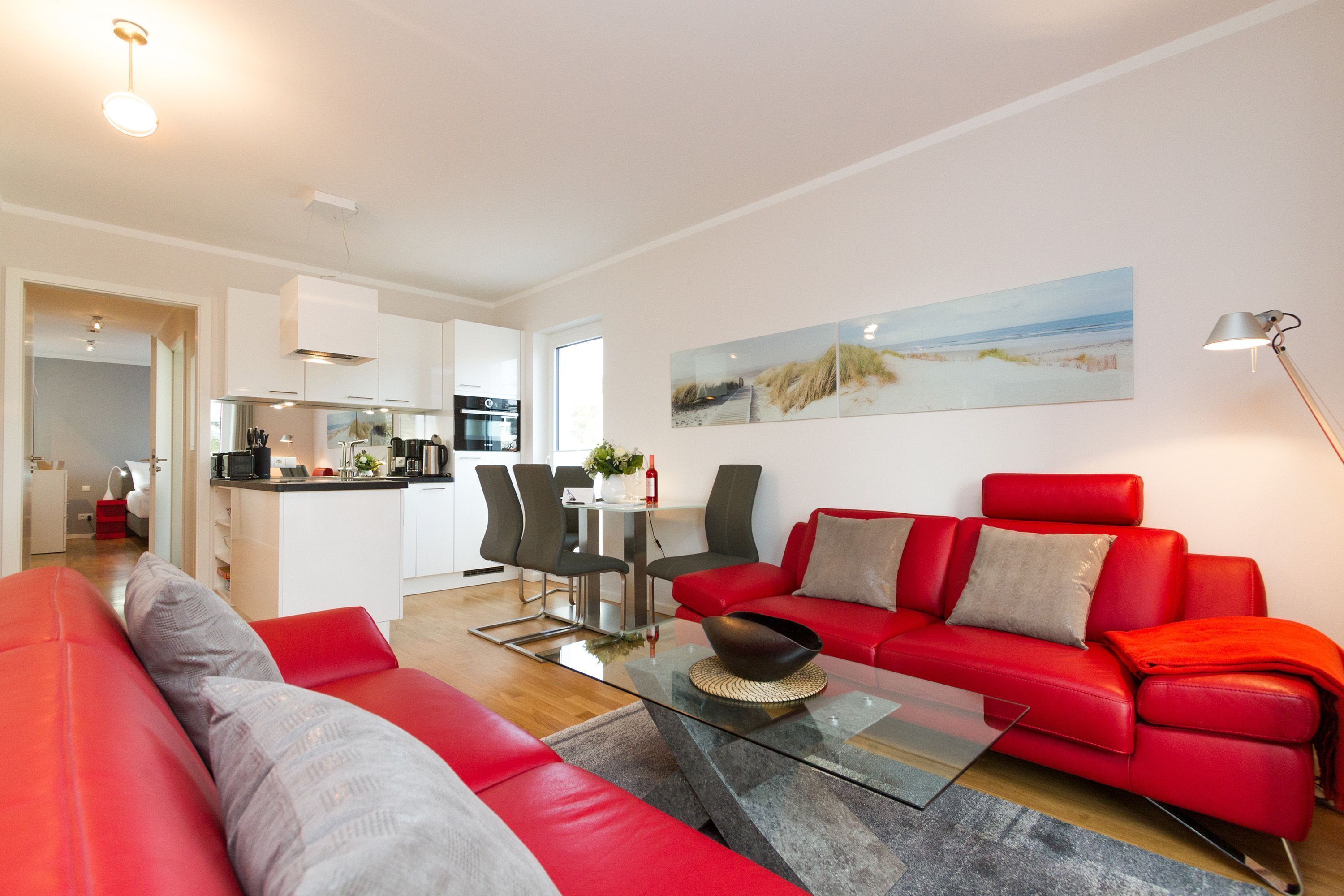 A modern living room with a red sofa, a glass coffee table, and a kitchen in the background.