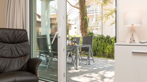 Ein modernes Zimmer mit Glastür zu einer Außenpatio. Das Zimmer bietet einen dunkelbraunen Ledersessel, einen weißen Schreibtisch mit Lampe sowie einen Blick auf einen Balkon mit Pflanzen und Gebäuden im Hintergrund.