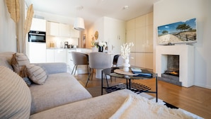 A modern living room with a sofa, a coffee table, and a fireplace. There is a kitchen in the background with cabinets and a stove. - Dünenresidenz Heringsdorf, App. 13 (Heringsdorf)