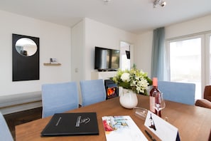 A hotel room with a dining table, a vase with flowers, a wine bottle, and a flat-screen TV mounted on the wall.