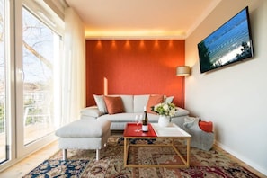 Un salon moderne avec un canapé, un pouf et une table basse. Une grande fenêtre laisse entrer la lumière naturelle et offre une vue sur l’extérieur.