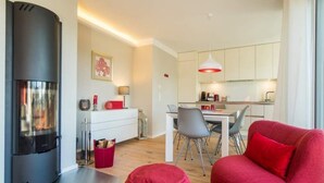 A modern living room with a fireplace, a dining area with a table and chairs, and a kitchen with white cabinets and pendant lighting. - Quartier am Fischerstrand, app. 4 (Heringsdorf)