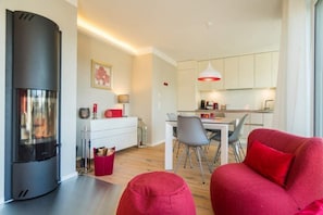 Un salon moderne avec une cheminée, un coin repas avec une table et des chaises, et une cuisine avec des meubles blancs et un éclairage suspendu.