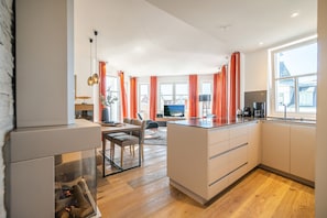 A modern kitchen with a dining area, a fireplace, and large windows with orange curtains. - Beachhouse Bansin, App. 23 (Heringsdorf)