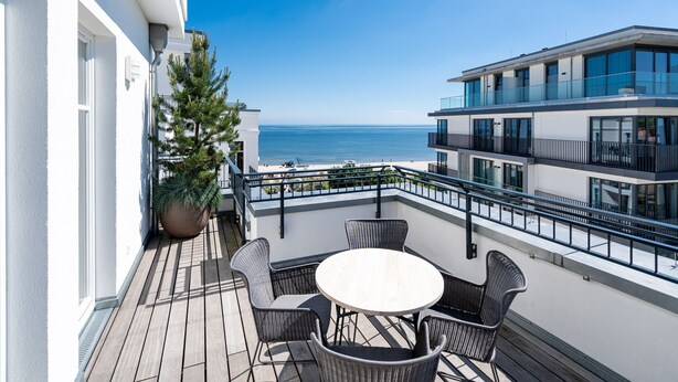 A balcony with a round table and wicker chairs, overlooking a beach and a modern building.