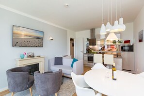 Een moderne woonkamer met een witte bank, grijze fauteuils en een eettafel met witte stoelen. Achterin is een haard te zien, en er is een keuken met moderne apparatuur zichtbaar door een open doorgang.