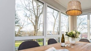 Ein Essbereich mit Holztisch, einer Weidenkorb-Pendelleuchte und großen Fenstern mit Blick auf Bäume und ein Gebäude.