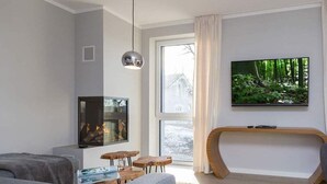 Ein modernes Wohnzimmer mit Kamin, einem an der Wand montierten Fernseher und einem großen Fenster mit Vorhängen.