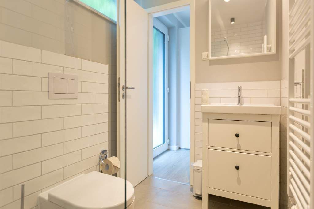 A modern bathroom with white tiles, a glass shower enclosure, a toilet, and a vanity with drawers.