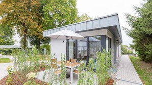 Un patio moderno al aire libre con una mesa y sillas, rodeado de plantas y un cerramiento de vidrio.