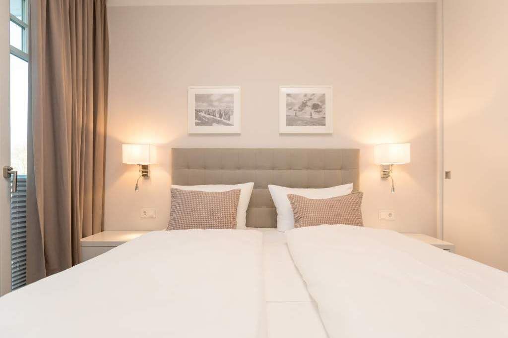 Habitación de hotel con dos camas, cabecero, lámparas fijadas a la pared y dos cuadros enmarcados.