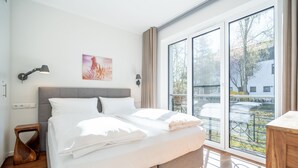 A modern bedroom with a large bed, a bedside table, a wall-mounted lamp, and a window with a view of the outdoors.