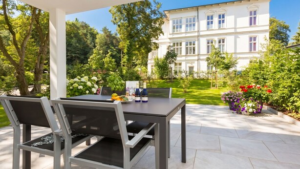 An outdoor patio with a table and chairs, a white building in the background, and a garden with various plants.