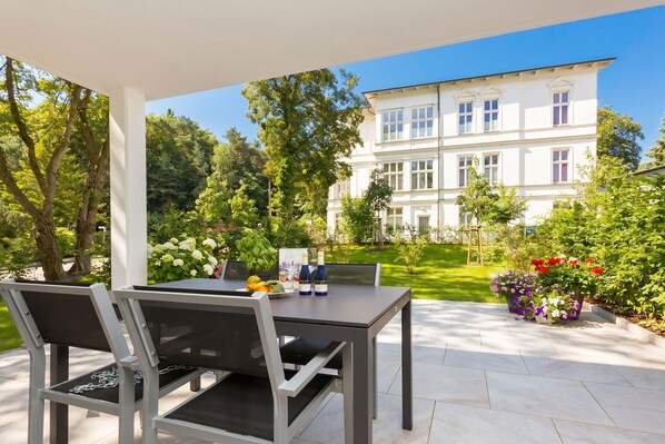An outdoor patio with a table and chairs, a white building in the background, and a garden with various plants.