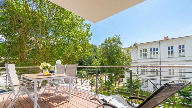 Una terraza de madera con mesa y sillas, una silla de descanso y vistas a un jardín y un edificio.