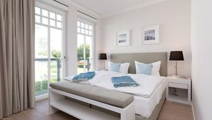 A bedroom with a large window, a bed with two pillows, a bedside table with a lamp, and a bench.