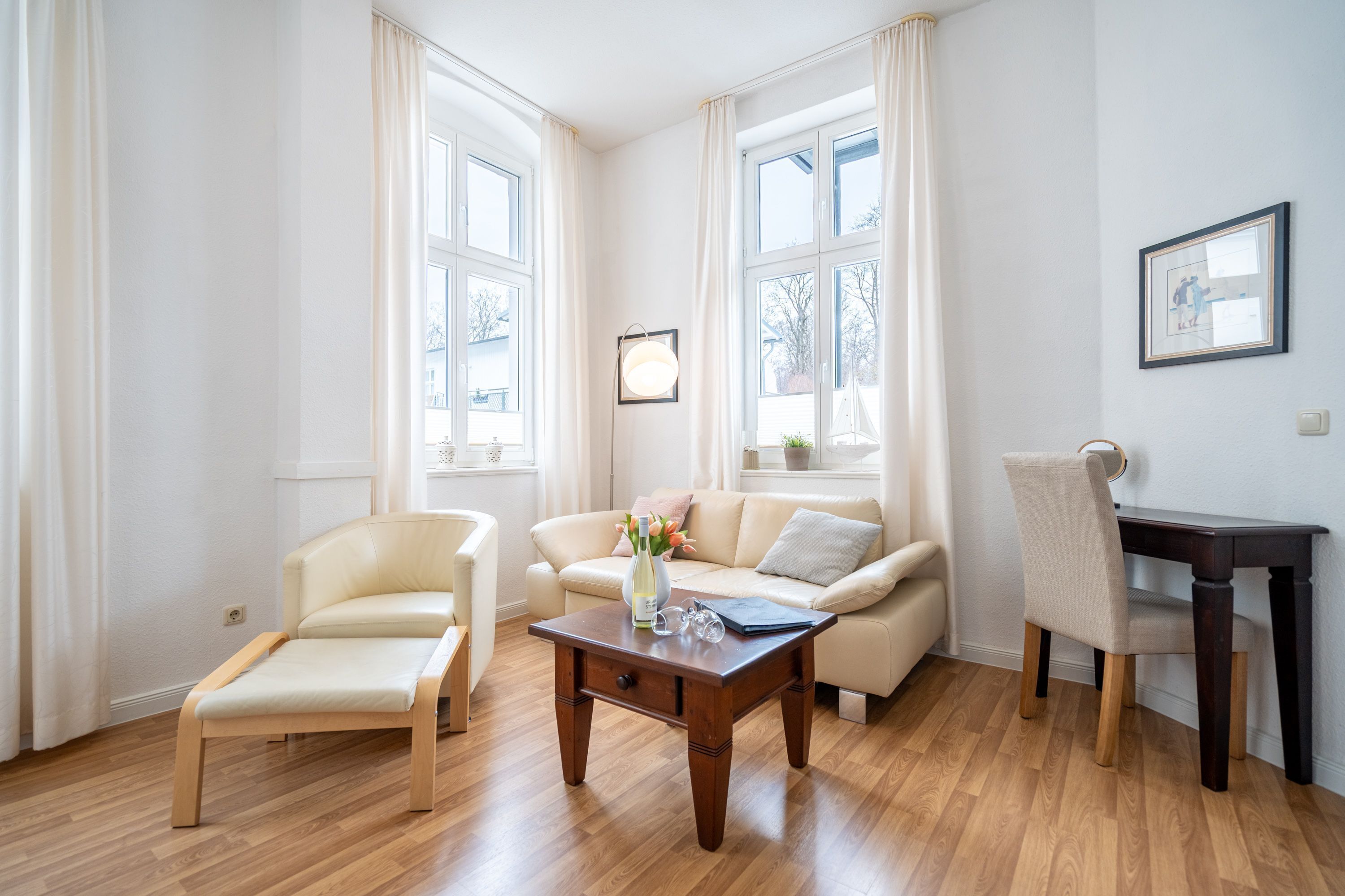 A room with a wooden floor, a sofa, a chair, a small table with a vase and glasses, and a framed picture on the wall.